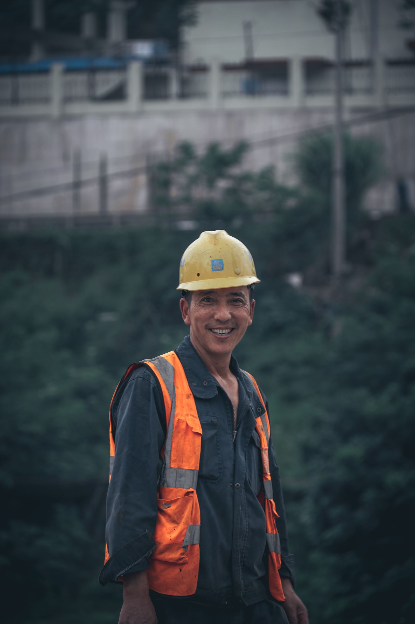 A smiling construction worker wearing safety gear stands outdoors in a work environment.