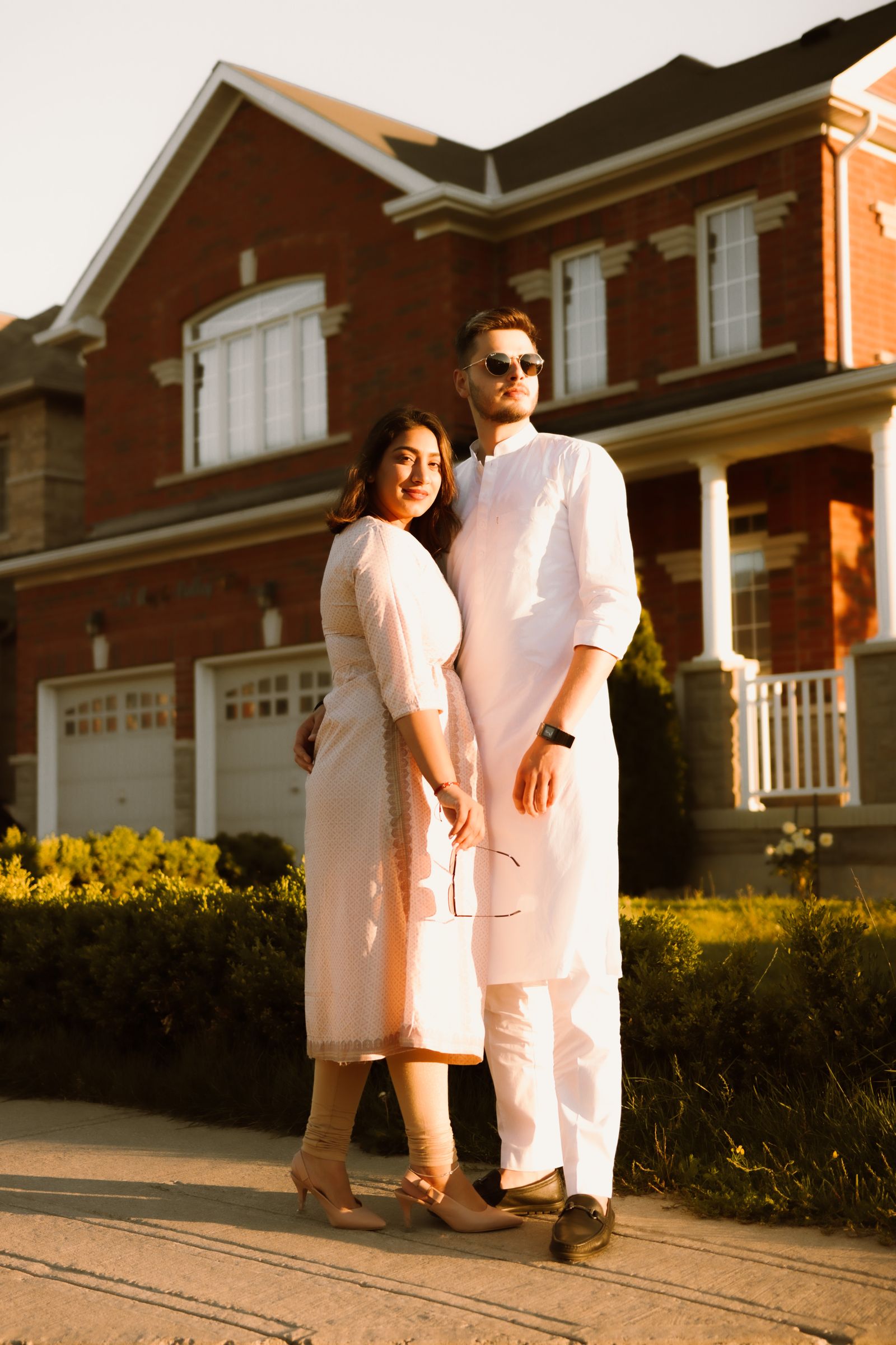 A fashionable couple in white attire posing outdoors in front of a luxurious brick house.