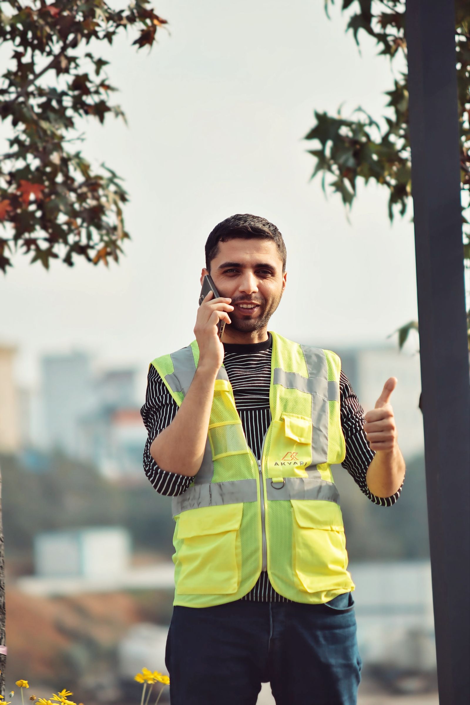 Young construction worker in a reflective vest talks on the phone and gives a thumbs up outdoors.
