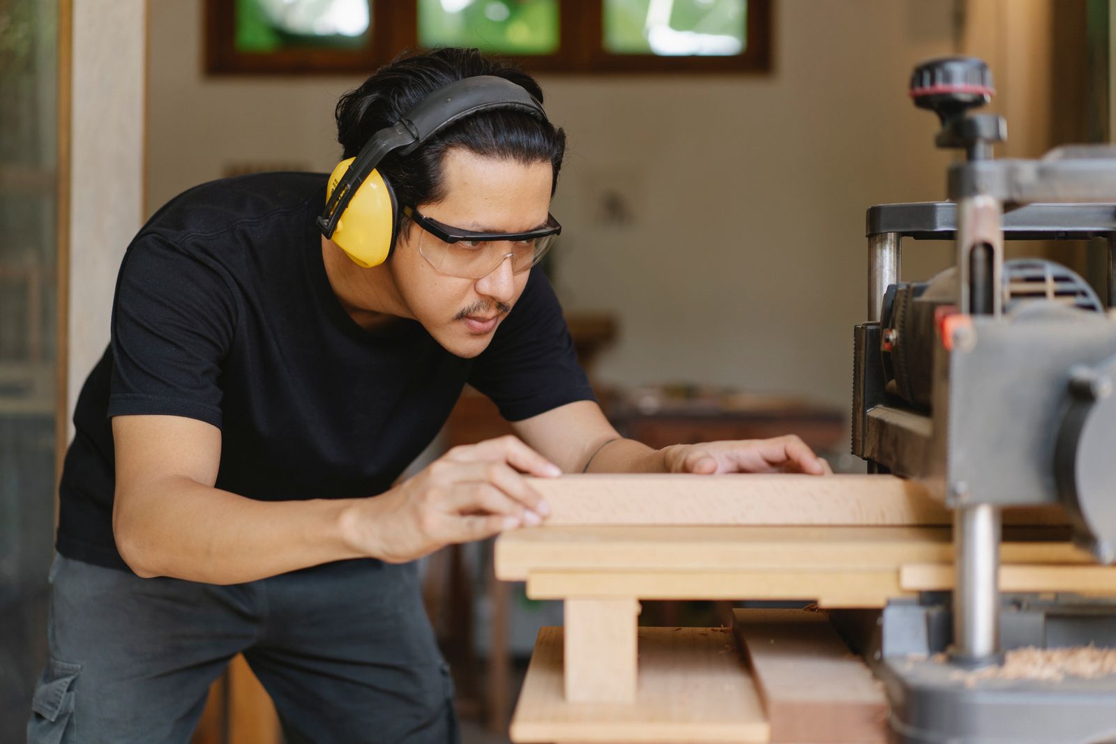 Young focused ethnic male carpenter in protective headphones and glasses flattening lumber on professional equipment in workroom